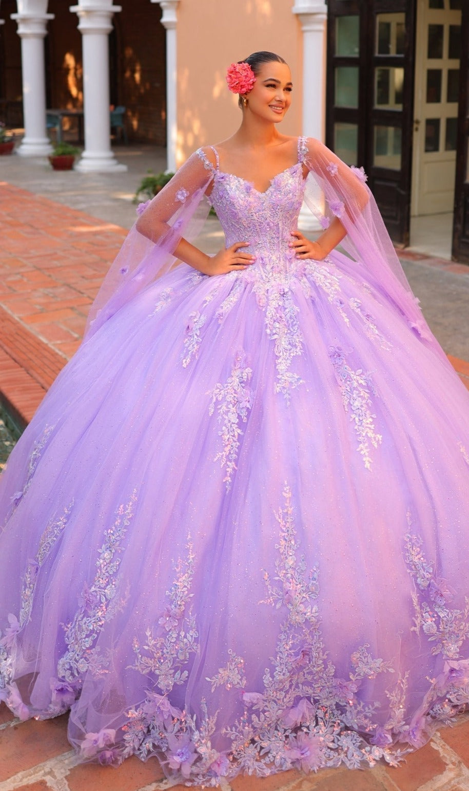 A woman stands outside in the Quinceanera Dress 54306 By Amarra?a voluminous lavender ball gown with floral embroidery, sheer details, hands on hips, and a pink flower in her hair.
